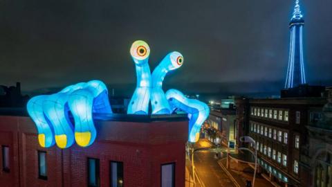 A huge blue inflatable monster on a rooftop in Blackpool on a dark night. Its right hand has three fingers with yellow nails holding onto the building and its head is made of two long pieces with googly eyes. Blackpool Tower can be seen in the distance to the right, lit up in blue