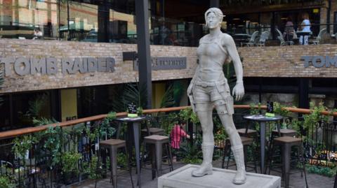 The nine foot (2.74m) statue of Lara Croft in a room with signs on the wall that says TOMB RAIDER