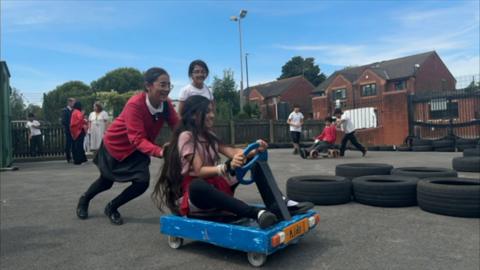 Three school girls in a playground smiling. One wears a red gingham dress sat on a blue go-kart. One wears a red cardigan, grey dress and white top and is pushing the go-kart around a stack of tyres. The third girl wears a white top, watching her friends.