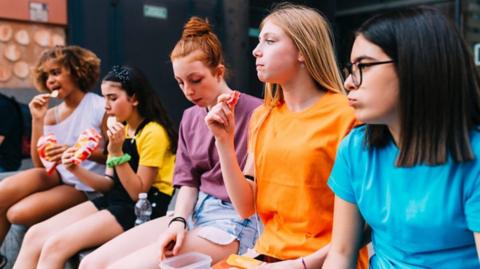 A group of teenagers sit alongside each other eating various snacks