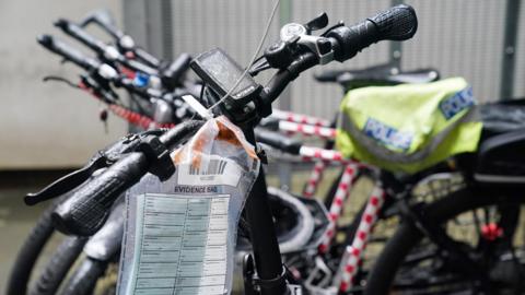 Bike handles are shown in the foreground with an evidence bag attached to them. In the background are a number of bikes, one has a police hi-vis jacket on the seat.