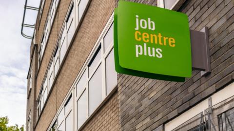 A green and white job centre plus sign on the outside of a red and grey brick modern building.