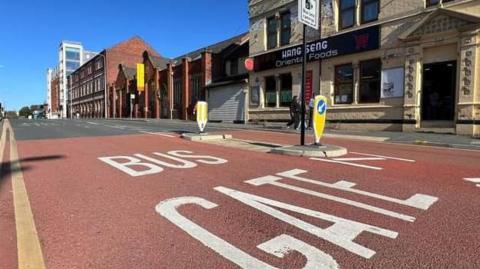 View of the words Bus Gate painted white on the road, with a sign next to it saying cameras are in force. Buildings are on the other side of the road including an Oriental food store.