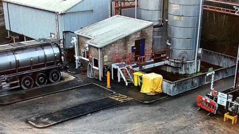 The outside of Futamura Chemical UK Ltd factory site. There are large metal cylinders, a large vehicle with a circular carrier and other factory equipment attached to different parts of the building. There are empty parking spots in front of the factory.