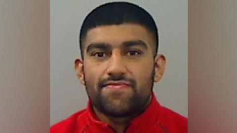 A mugshot of Ameer Khalile who has dark hair, closely cropped and has a closely cropped beard and moustache. He is wearing a red top.