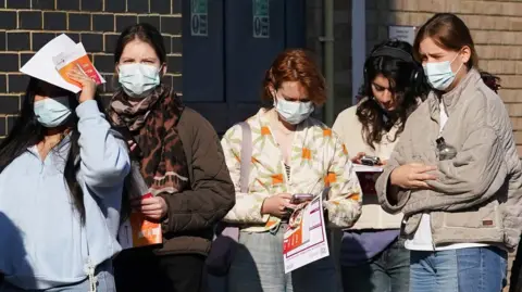 Crowd of five girls standing outside building, four wear facemasks and some hold leaflets in their hands