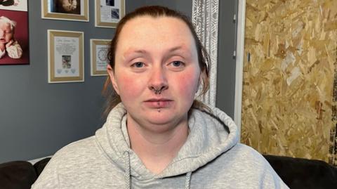 Chloe Rutherford is seen standing in her living room. She has brown hair and a number of nose and lip piercings and is wearing a grey hoodie. A number of family portraits are visible on the wall in the background. A large plywood wooden sheet covers the window that was smashed.