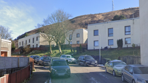 A street with cars parked on either side and two storey white houses with flat roofs. A grassy hill can be seen behind the houses