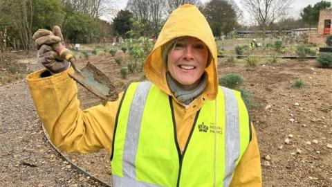 Sonja, wearing a grey hat and scarf underneath a yellow raincoat and Royal Parks hi vis waistcoat, holds up a muddied trowel and smiles