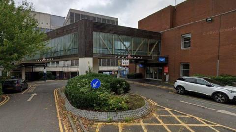 A wooden bridge with large glass windows connecting two hospital buildings, over a car park