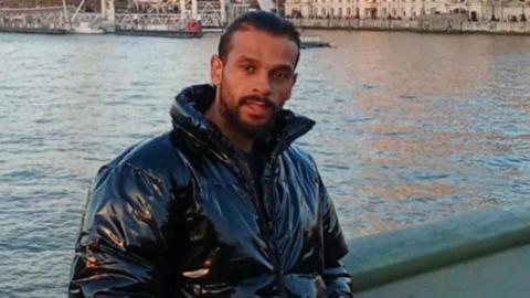 An Asian man in London. He has his hair tied back and a beard. There is the River Thames behind him. He is wearing a black puffer jacket. It looks like the sun is going down. He is near the London Eye.