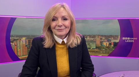 The Mayor, Tracy Brabin is pictured in a Radio Leeds studio. She is wearing a yellow jumper and black suit jacket. An image of Leeds is behind her in a screen.