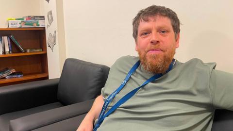 A man in a green t-shirt and blue lanyard sat on a black sofa. He has short dark hair and a ginger beard. There is a another matching black armchair and a bookshelf beside him.