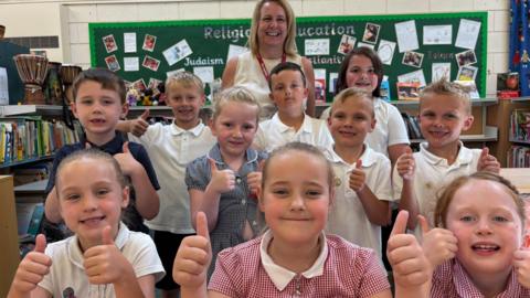 Lots of smiley school children huddle together with their thumbs up. A proud teacher stands behind them