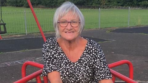 A woman, with grey hair and wearing a white and black stop, smiles at the camera while in a children's play park 