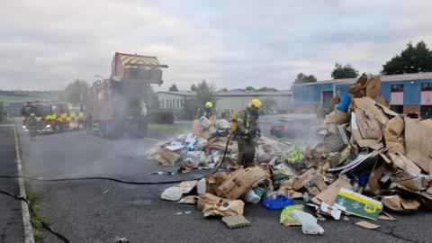 Two firefighters pour water from hoses on a large pile of cardboard and other recycled material while dealing with a fire. Smoke is coming from the pile. The lorry the items were in is parked near the firefighters. A fire engine is next to the lorry.