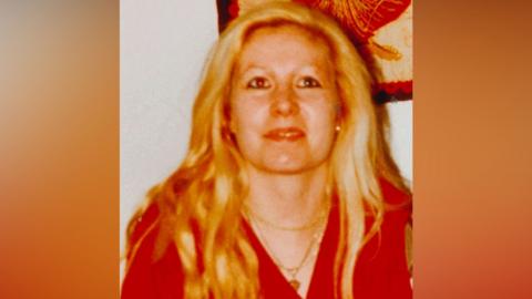 An old, heavily saturated photograph from the early 1990s of a woman with long blonde hair, who is wearing a stack of gold necklaces and a v-neck red top. She is looking above the shot and is slightly smiling.