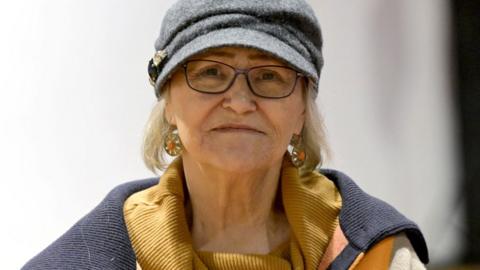 Pamela Hemphill wears a golden scalf and a grey hat as she looks directly at the camera in a portrait photograph