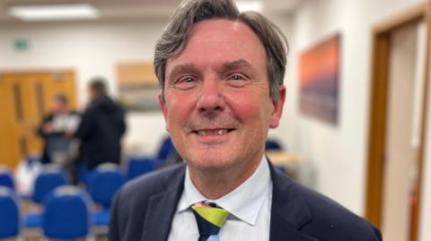 Deputy Andy Sloan - A man with coifed black and grey hair smiling at the camera. He is wearing a blue suit and a multi coloured tie on top of a white shirt. 