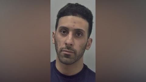 A police mugshot of a man wearing a dark blue top. He has black hair and is looking directly at the camera.