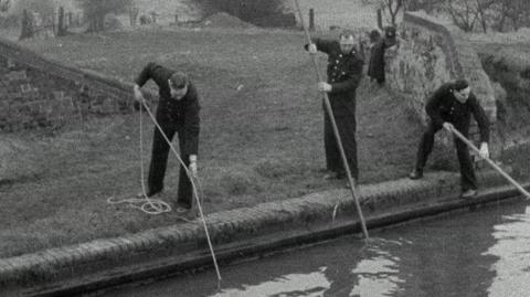 A still from black-and-white news footage dating back to the original investigation. It shows three uniformed police officers searching a waterway from the bank with poles and ropes.