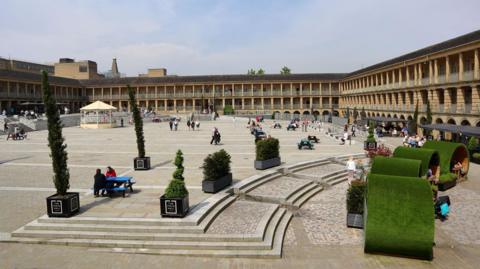 The exterior of a large building set in a square shape with a central courtyard. There are people sitting in the courtyard with greenery dotted around. 