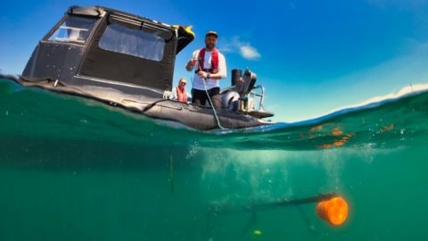 An underwater camera capturing below sea level and the surface. To the left is a small boat with a man stood at the front, with wire of sorts attached to the underwater camera. Below the surface is a piece of equipment with an orange float on the right.