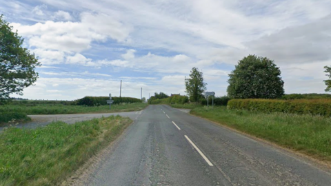 A junction on a quiet country road, lined on both sides with long grass and hedges