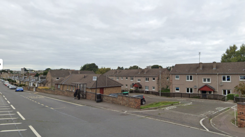 A road lined with brown houses. There is a bus stop and some parked cars