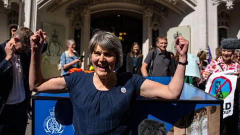 A woman wearing a dark blue shirt is standing outside a Supreme Court. There are campaigners holding banners standing behind her.