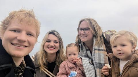 Ed Sheeran poses for a selfie with Amanda Jane, her friend, and their children. Ed has ginger hair and is smiling into the camera. Amanda is wearing glasses and a scarf while her female friend has blonde hair. The two children pictured are each holding a piece of food and staring into the camera. 