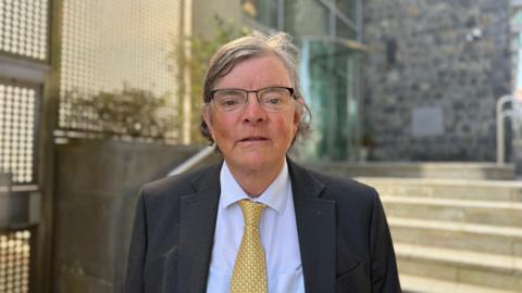 Deputy Charles Parkinson - A late middle aged man with long brown and grey hair. He is wearing glasses. He has a blue shirt, yellow tie and dark grey blazer on.