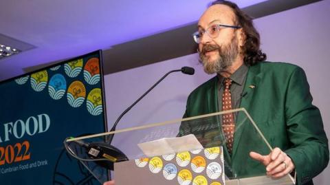 Myers is standing in front of a stand and microphone next to the blue Cumbria Food Awards sign. He is looking side on and is wearing a shiny green jacket, grey shirt and brown tie. His moustache is curled up at the ends. 