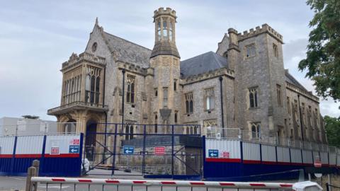 A historic building made of beige stones with big windows and towers. It is surrounded by a fence that suggests it's a building site.