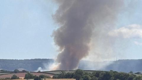 Thick black smoke billows over fields in Devon. It is rising into the air.