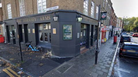 The exterior of a pub called the Hornsey Arms from the road. The pub is on the corner of a street, and the picture is taken from the road looking towards the outer corner. The pub's exterior is dark grey, with a green sign on the exterior wall. On the right, there is a road lined with parked cars. The road continues to the left. Above the pub, there is a street sign reading St Mary's Road.