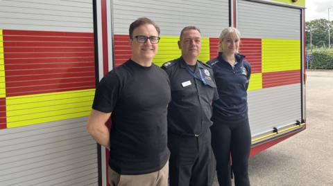 Professor Athanassios Bissas, Chris Winter and Anna Ferguson standing against a fire truck wearing firefighter's navy shirt.