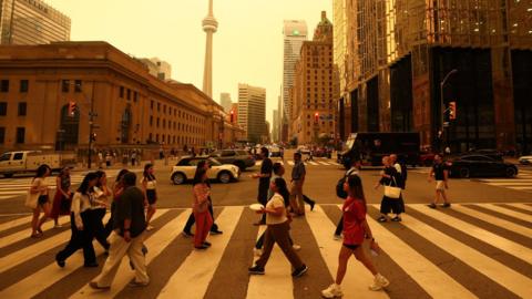 People walk on a crossing in a road downtown Toronto. There are buildings in the background and sky is orange from smoke.