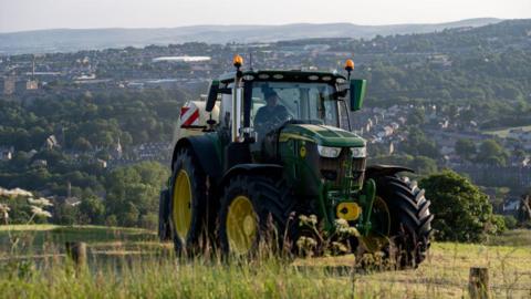 A green tractor drives through a field, with a view of a valley in the background.