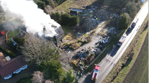 A drone image of smoke billowing from a large house, with scrub and waste land to the right, and housing on the other. Fire engines are parked on the country lane outside, adjoined by a green field. 