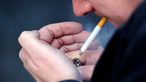 A man has a cigarette in his mouth and is cupping his hands around his red lighter. The cigarette is lit and giving off smoke