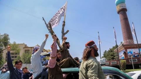 Two men brandishing weapons ride on top of a green car with others around them raising fists