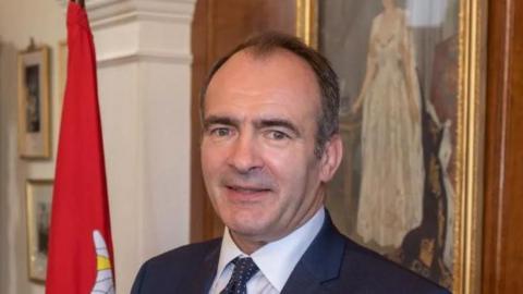 Alfred Cannan smiling. He is wearing a dark suit and tie with a white shirt, and is standing next to a Manx flag. There is a portrait of Queen Elizabeth II on the wall behind him.