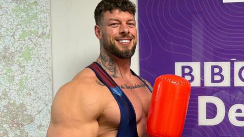 Giant - real name Jamie Christian-Johal from Ilkeston stood with a inflatable pugil stick in front of a sign which reads: BBC RADIO Derby. He is wearing a lycra body suit flexing his right bicep which is exceptionally large.  