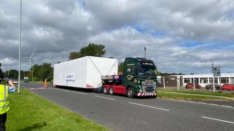 A building pod on a trailer, with workers in hi vis either side of the vehicle.