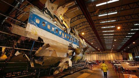 The wreckage of the plane has been reconstructed in a warehouse. It is white with a blue stripe and black lettering, and it has missing and damaged sections. A person in a high viz jacket is walking next to the wreckage.