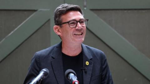 Andy Burnham is standing in front of a green background. He is a white man with dark hair and is wearing glasses, a black t-shirt and blue jacket with a yellow bee pin badge.