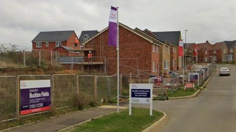 Homes that are partially built on a housing estate, made from red brick, showing one car on the street, a number of signs and a flag pole with a purple flag on it. There is some scaffolding on the home nearest the camera.