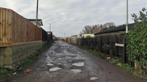 A muddy path with puddles has wooden fencing on either side as well as a white sign that says 'The Laurels'. The tops of mobile homes can be seen above the fences. 