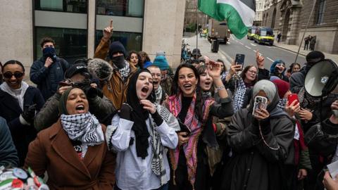 A group of protesters stand on a street corner in London. Some of them are shouting and smiling with their hands in the air. Many are holding phones up, one is holding a camera with a fluffy microphone and one has a megaphone.
